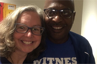 [ai] A woman with glasses smiling next to a man wearing glasses and a blue shirt, both looking at the camera. The background features colorful artwork.